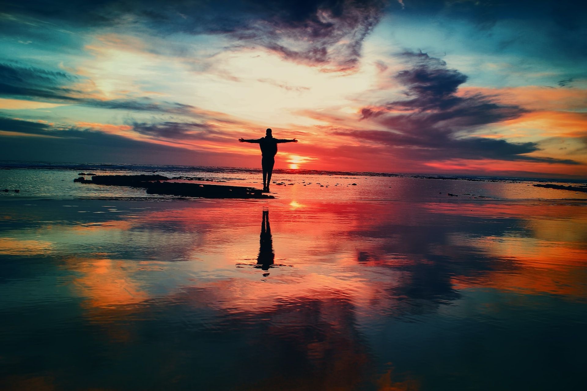 Vor einem wolkenverhangenen roten Sonnenuntergang steht eine Person mit ausgebreiteten Armen am Strand.